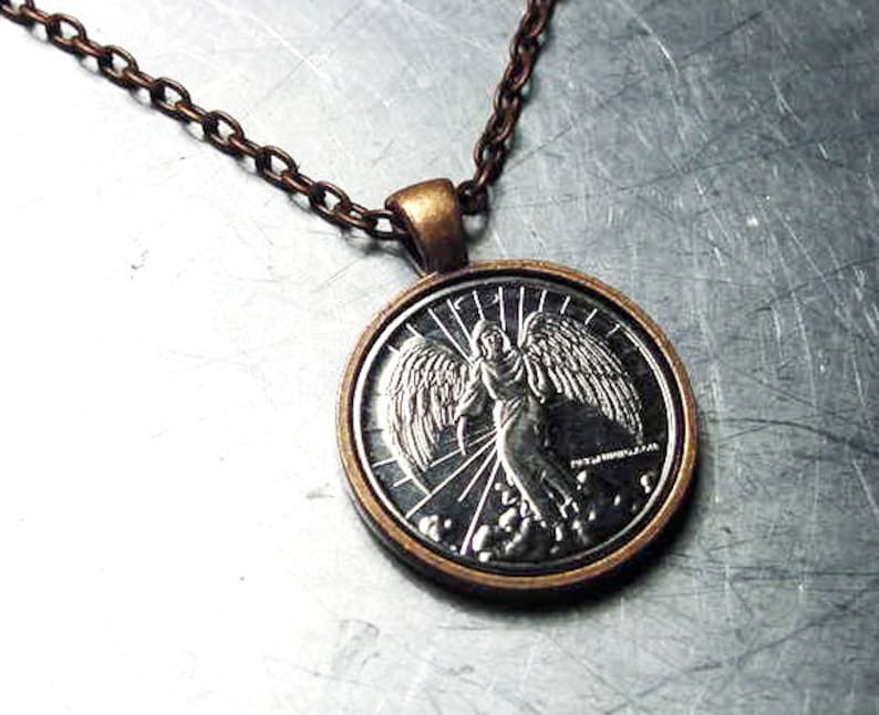 May include: A silver pendant necklace with a round, silver coin featuring a detailed image of an angel with wings spread wide. The coin is surrounded by a copper-colored frame.