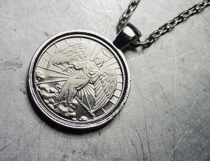 May include: Silver pendant necklace featuring a detailed image of an angel with wings spread, surrounded by a halo and rays of light. The pendant is attached to a silver chain.