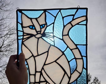 May include: A rectangular stained glass panel featuring a white cat with bright blue eyes against a light blue background. The cat is composed of multiple white glass segments outlined in black lead, with a long tail curving upward.