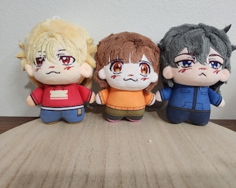 Op de afbeelding: Drie pluche poppen met geborduurde gezichtskenmerken en haar. De linkerpop heeft blond haar en een rode hoodie, de middelste pop heeft bruin haar en een oranje trui, en de rechterpop heeft donkergrijs haar en een blauw jasje.