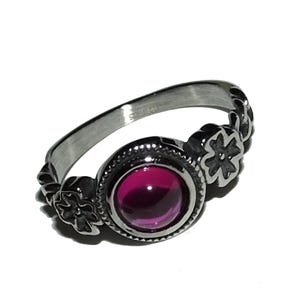 May include: A silver-colored ring with a large, round, deep pink gemstone at the center. The ring features floral designs on the sides and around the gemstone. The band is smooth and polished, with a subtle texture. The ring is set against a white background.