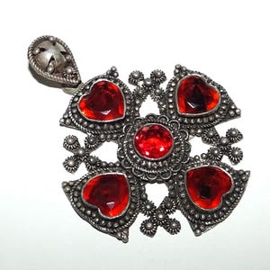 May include: A silver pendant with a cross design featuring five red heart-shaped stones. The pendant has a detailed filigree design.