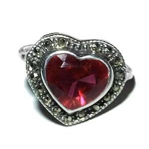 May include: A silver ring with a heart-shaped, faceted, deep red gemstone. The gemstone is surrounded by a border of small, dark, round stones. The ring is set against a white background.