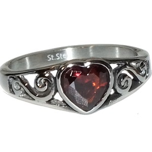 May include: A silver ring with a heart-shaped red gemstone set in a delicate filigree design. The ring is engraved with the words "St. Steel".