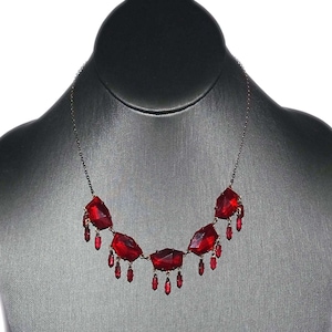 May include: A silver-toned necklace featuring large, faceted, red gemstone pendants. Each pendant has smaller, dangling red teardrop-shaped gems. The necklace is displayed on a gray mannequin bust.