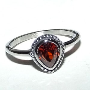 May include: A silver ring with a red pear-shaped gemstone set in a decorative silver bezel. The ring is marked "St. Steel" on the inside band.