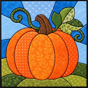 May include: A square fabric quilt block featuring a central orange pumpkin with segments in varying patterns of dots and leaves. The pumpkin has a brown stem, green curly vines, and green leaves, set against a blue and white geometric background.
