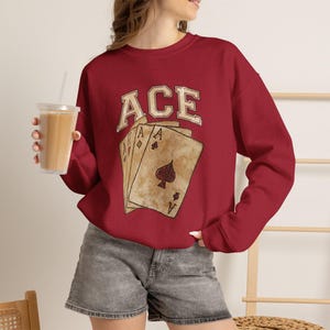 May include: A dark red crewneck sweatshirt featuring a graphic print of three playing cards with the word ACE arched above in a collegiate font. The cards are depicted with a vintage, distressed texture and a prominent spade symbol.