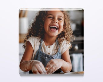 May include: A square photo print featuring a smiling child with curly hair wearing a white shirt and a blue denim apron. The child has flour on their hands and is engaged in a baking activity, captured in a bright, indoor kitchen setting.