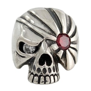 May include: A silver-toned skull ring featuring a clear, round-cut gemstone in the left eye socket and a deep red, round-cut gemstone in the right eye socket, surrounded by a radiating sunburst pattern etched into the metal surface.