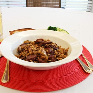 May include: A white bowl filled with a brown rice dish with sausage, mushrooms, and herbs, sitting on a red placemat with two silver forks. A glass of beer and a plate with bread and green vegetables are in the background.