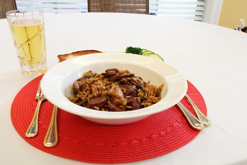 May include: A white bowl filled with a brown rice dish with sausage, mushrooms, and herbs, sitting on a red placemat with two silver forks. A glass of beer and a plate with bread and green vegetables are in the background.