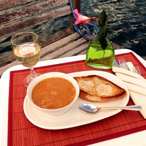 May include: A white plate with a bowl of soup, a slice of toasted bread, a spoon, and a glass of white wine. The plate is on a red and brown placemat. The table is white and there is a green glass vase with a pink flower in the background.