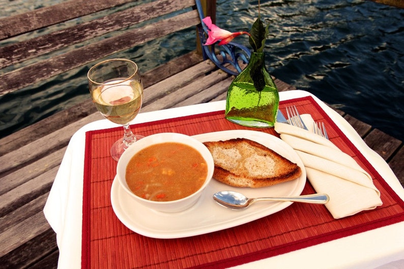 May include: A white plate with a bowl of soup, a slice of toasted bread, a spoon, and a glass of white wine. The plate is on a red and brown placemat. The table is white and there is a green glass vase with a pink flower in the background.