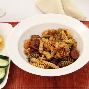 May include: A white bowl filled with a pasta dish with green and brown pasta, shrimp, sausage, and a brown sauce. The bowl is on a brown bamboo placemat with a white napkin and a glass of wine in the background.