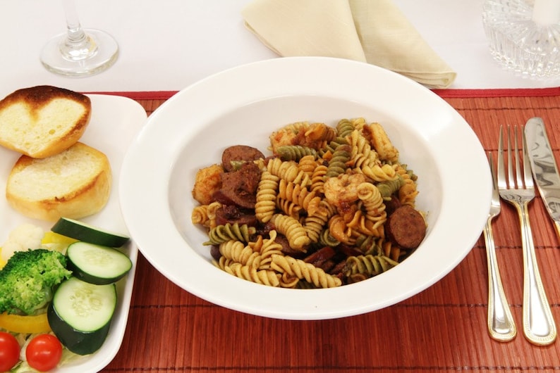 May include: A white bowl filled with a pasta dish with green and brown pasta, shrimp, sausage, and a brown sauce. The bowl is on a brown bamboo placemat with a white napkin and a glass of wine in the background.