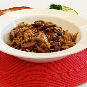 May include: A white bowl filled with jambalaya, a rice dish with sausage and meat. The bowl sits on a red placemat, with a glass of liquid, a slice of toast, and silverware.