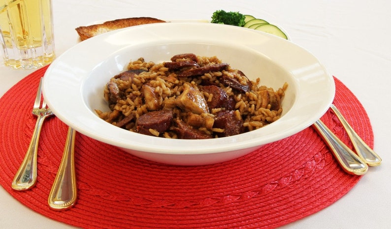 May include: A white bowl filled with jambalaya, a rice dish with sausage and meat. The bowl sits on a red placemat, with a glass of liquid, a slice of toast, and silverware.