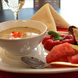 May include: A white bowl of soup with red pepper garnish sits on a white plate, alongside a spoon, red bell pepper, watermelon, avocado, and toasted bread. A glass of white wine and a folded napkin are in the background.
