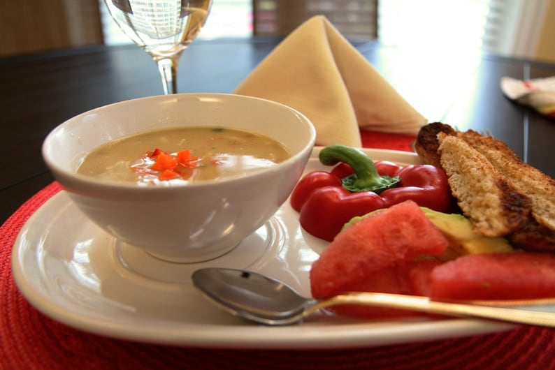 May include: A white bowl of soup with red pepper garnish sits on a white plate, alongside a spoon, red bell pepper, watermelon, avocado, and toasted bread. A glass of white wine and a folded napkin are in the background.
