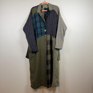 May include: A patchwork coat with a variety of fabrics and patterns. The coat features a mix of green, blue, gray, and plaid fabrics. It has a collared neckline, button closures, and long sleeves. The coat is hanging on a pink hanger.