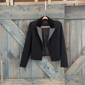 May include: A black blazer with a faux fur collar and a white and black dotted pattern on the lapels. The jacket is hanging on a pink hanger against a blue wooden door.