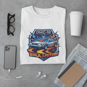 Könnte beinhalten: Ein weißes T-Shirt mit einer Grafik eines DMC-12 Autos und dem Text BACK TO THE FUTURE. Das Design zeigt bunte Blitze, einen Gitterhintergrund und orangefarbene Straßenlinien, alles innerhalb einer blau umrandeten Schildform.