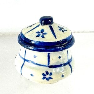May include: A small, ceramic jar with a lid. The jar is off-white with blue floral and geometric patterns. The lid and rim are a darker blue. The jar is likely used for storage or decoration.