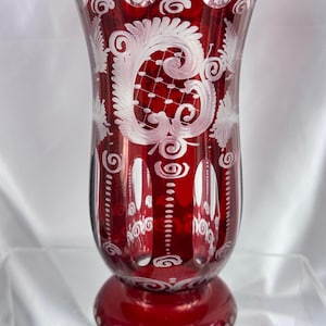 May include: A ruby red glass vase with white etched floral and scroll designs. The vase has a wide, flared top and a rounded base. The etched patterns include swirls, dots, and a large central design. The vase is likely used for decorative purposes.