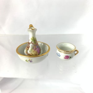 May include: A miniature porcelain set featuring a white pitcher with a gold rim and a floral design, and a white chamber pot with a gold rim and a pink rose design. The chamber pot has the word "Nuit" written on it.
