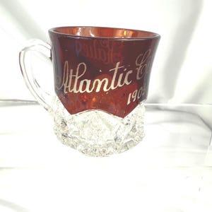 以下が含まれることがあります： 赤いガラス製の記念マグカップで、透明なガラスの底があります。マグカップには持ち手があり、金色の文字で「アトランティックシティ 1903」と書かれています。
