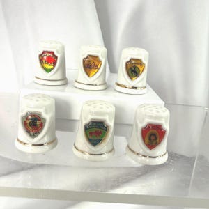 May include: A collection of white porcelain thimbles with gold trim. Each thimble features a different colorful emblem, including images of landscapes and text. The thimbles are arranged on a clear surface.