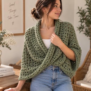 May include: A sage green crochet shrug featuring an openwork stitch pattern, a wide collar, and a wrap-front design. The garment is worn over a white top and blue denim jeans, showcasing a textured, lightweight knit construction.