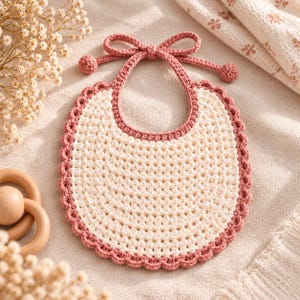 Puede incluir: Un babero de bebé tejido a ganchillo con un centro color crema de punto texturizado, bordeado por un ribete festoneado en rosa empolvado. El babero incluye lazos de ganchillo a juego en rosa empolvado, rematados con una pequeña bola.