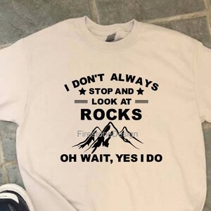 May include: A beige t-shirt with black text that reads "I DON'T ALWAYS STOP AND LOOK AT ROCKS OH WAIT, YES I DO." The shirt has a mountain graphic. A pair of black Adidas sneakers with white stripes are next to the shirt.