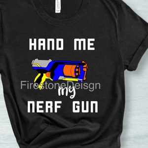 Puede incluir: Camiseta negra con texto blanco que dice "Hand Me My Nerf Gun". Una imagen de dibujos animados de una pistola Nerf azul y amarilla está en el centro de la camiseta.