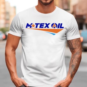 Peut inclure: T-shirt blanc avec l'inscription "M TEX OIL" en bleu et orange. Le design comprend une étoile et une flamme. Le t-shirt est à manches courtes et porté par une personne.