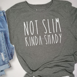 May include: A gray t-shirt with the text "NOT SLIM KINDA SHADY" in white, paired with folded blue denim jeans with distressed details. The t-shirt is made of a soft material and is suitable for casual wear.