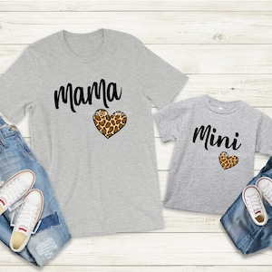 May include: Two gray t-shirts, one adult and one child size, with the words "Mama" and "Mini" in black script. Each shirt features a leopard print heart. Blue jeans and white sneakers are also in the image.