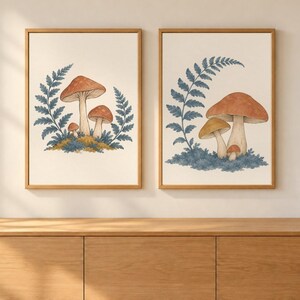 May include: Two framed wall art prints featuring botanical illustrations of mushrooms and fern fronds. Each print displays clusters of brown and tan mushrooms nestled in blue and green mossy patches, accented by detailed blue fern leaves on a cream background.