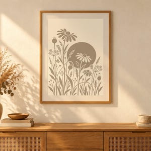 May include: A framed wall art print featuring a beige background with a taupe-colored graphic illustration of various wildflowers, grasses, and a large solid circle representing the sun, all enclosed in a simple light wood frame.