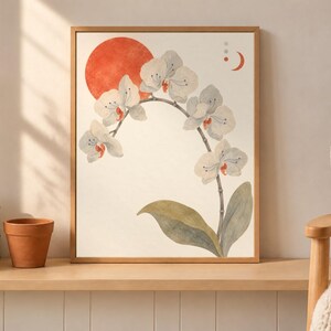 May include: A framed wall art print featuring a white orchid branch with green leaves against a cream background. A large solid orange circle sits behind the flowers, accompanied by a small orange crescent moon and three vertical dots in the upper right.