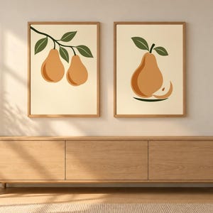 May include: Two framed wall art prints featuring stylized pear illustrations. The left print shows two pears on a branch with green leaves. The right print displays a single pear with a slice and green leaves. Both have light cream backgrounds.