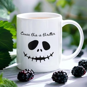 Puede incluir: Una taza de cerámica blanca con un gráfico negro de un rostro esquelético con ojos huecos y boca cosida. Sobre el rostro, el texto "Cause this is thriller" está impreso en una fuente cursiva negra de estilo manuscrito.