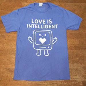 Puede incluir: Una camiseta azul con el texto "LOVE IS INTELLIGENT" sobre un personaje de dibujos animados de computadora blanco con una pantalla en forma de corazón. El personaje tiene brazos, piernas y una cara sonriente.