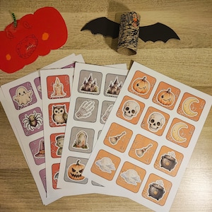 May include: Four sheets of Halloween-themed stickers featuring ghosts, spiders, owls, pumpkins, skulls, moons, and cauldrons. A red paper pumpkin cutout and a cardboard tube bat with black paper wings are positioned above the sticker sheets.