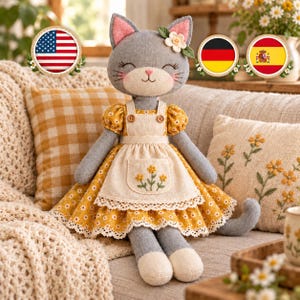 May include: A plush gray cat doll with embroidered facial features and pink inner ears. The cat wears a yellow dress with a white daisy pattern, a cream-colored apron with embroidered yellow flowers, and a small white flower on its head.