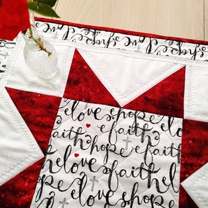 Pode incluir: Um corredor de mesa acolchoado branco e vermelho com um design de estrela. O corredor de mesa tem letras pretas que dizem "love, hope, faith" repetidas em todo.