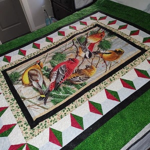 Peut inclure: Une courtepointe verte, rouge et blanche avec une bordure noire. Le centre de la courtepointe présente une illustration colorée d'oiseaux dans une scène d'hiver.