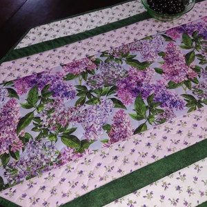 May include: A quilted table runner with a lilac floral print in shades of purple and green. The runner has a light blue background and is trimmed with green fabric.